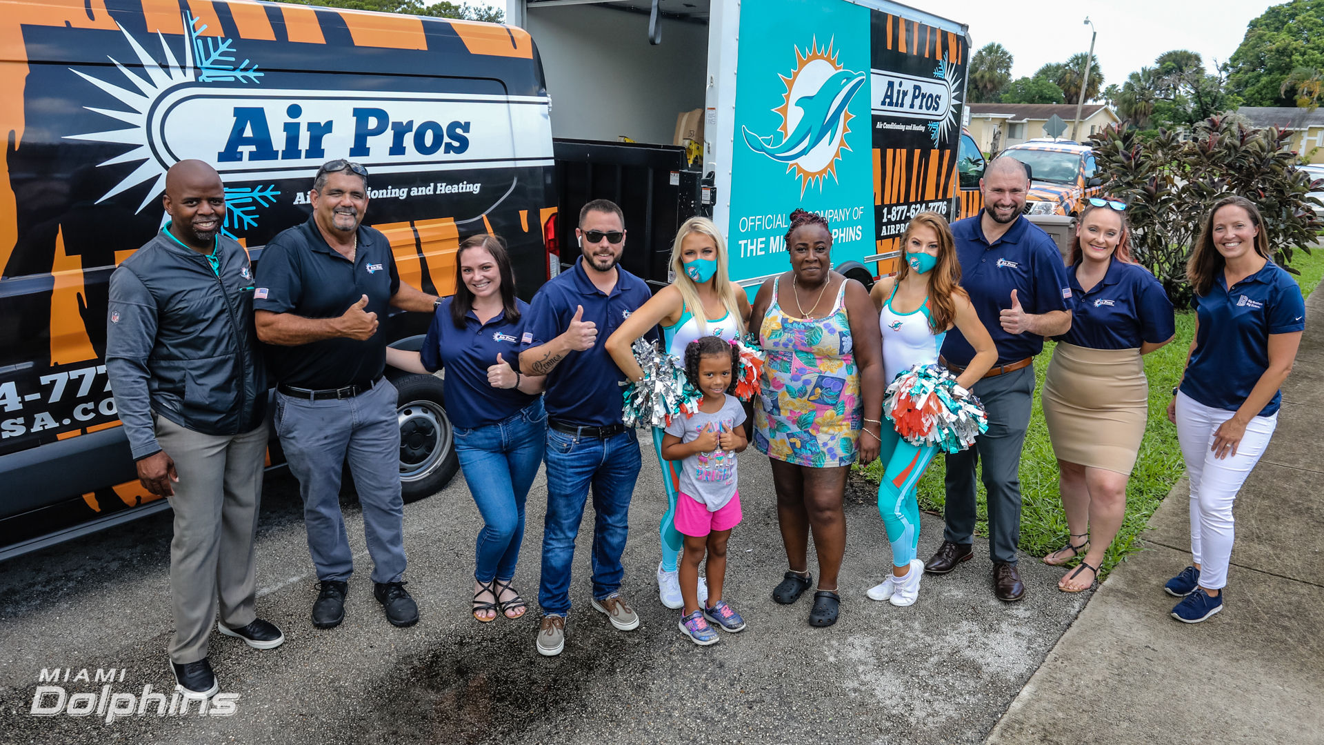 A group of people, including Miami Dolphins cheerleaders, Air Pros staff, and Big Brothers Big Sisters Broward members, pose together outdoors in front of branded vehicles, some giving thumbs up.