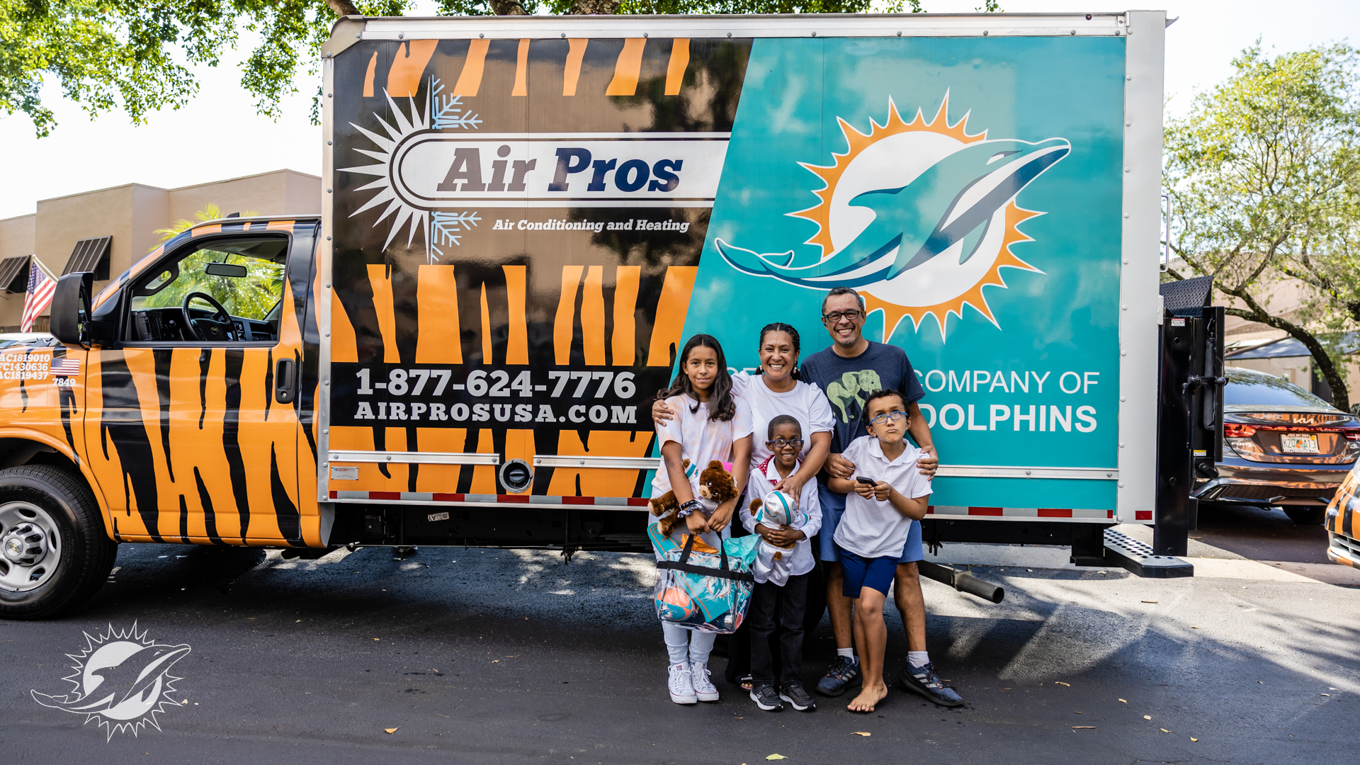 A family of five stands in front of an Air Pros USA truck with a Miami Dolphins logo, posing for a photo outdoors on a sunny day—possibly celebrating one of the company’s free A/C units giveaways.
