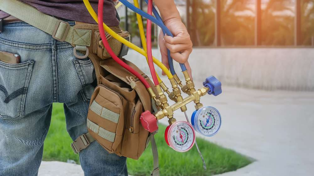 A person with a tool belt holds a set of HVAC manifold gauges with red, yellow, and blue hoses, standing outdoors near a building during central AC maintenance.