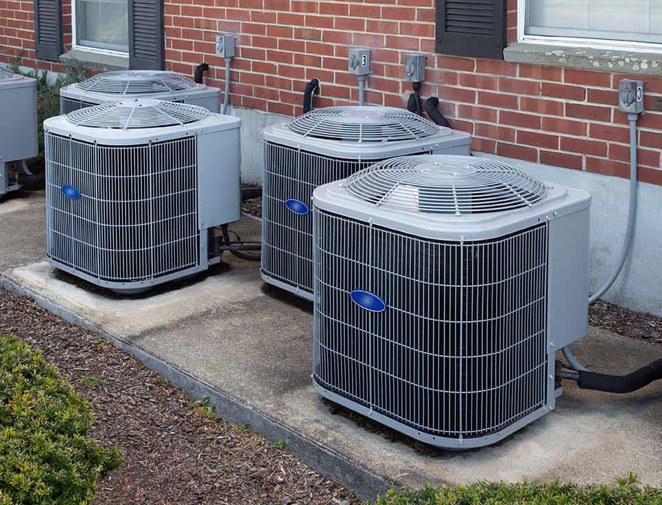 Three Central AC units are installed on concrete pads next to a brick building, with visible electrical connections, ensuring the space is cooled efficiently.