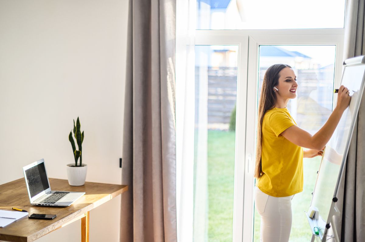 A woman in a yellow shirt writes on a flip chart near a glass door, with a desk holding a laptop, notebook, and plant from Air Pros in the background—promoting the benefits of a free HVAC UV light for cleaner air.