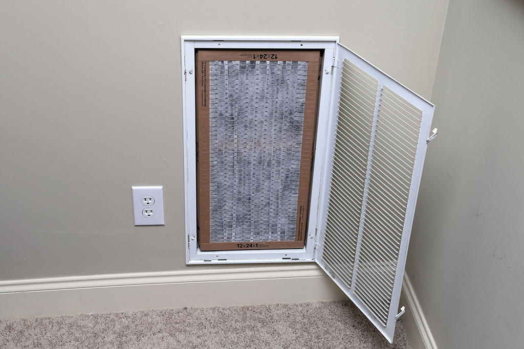 An air return vent with its cover open, revealing a rectangular AC filter installed inside, located on a beige wall near an electrical outlet and carpeted floor—perfect for anyone focused on AC maintenance.