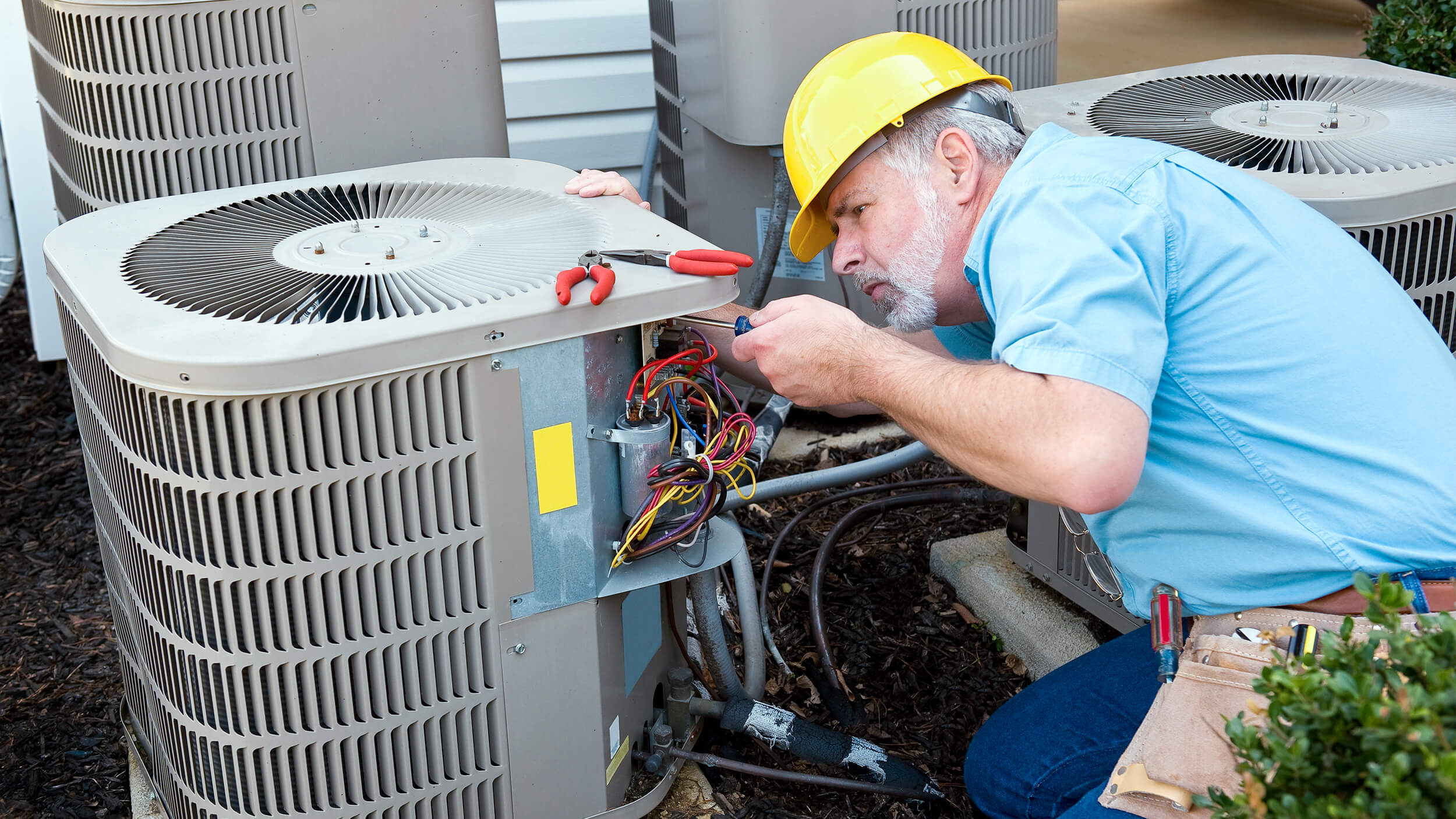 An HVAC technician wearing a yellow hard hat repairs wiring inside a central AC unit with a screwdriver, helping to extend AC lifespan and ensure reliable cooling.