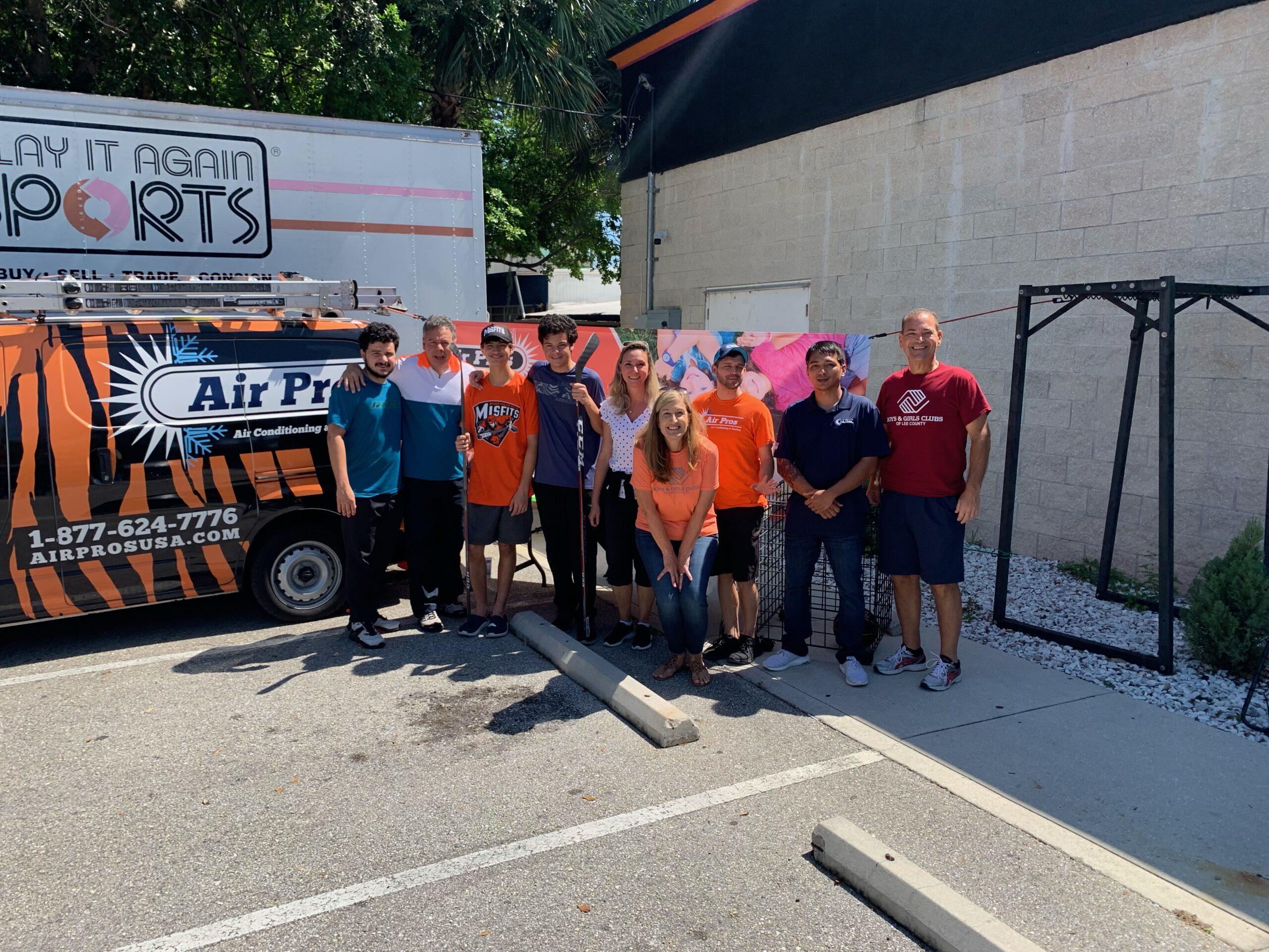 A group of ten people stands together in a parking lot near an Air Pros USA van and a Play It Again Sports Fort Myers truck on a sunny day.
