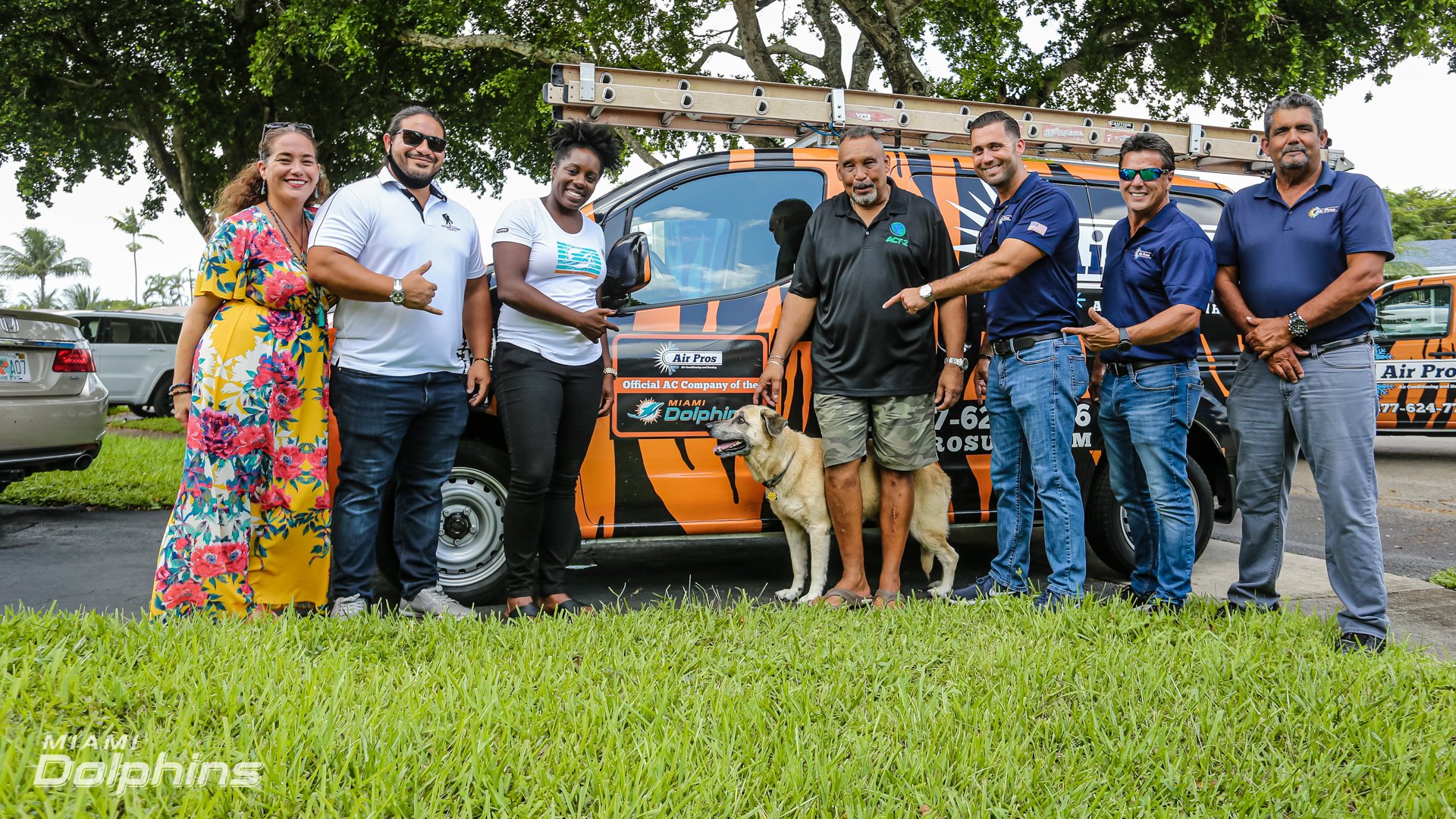 A group of seven adults and a dog stand in front of an Air Pros USA van with a ladder on top. The Miami Dolphins logo is visible on the van and in the corner, highlighting their partnership.