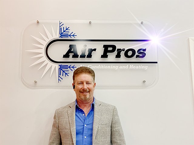 A man in a gray plaid jacket and blue shirt stands in front of an "Air Pros" air conditioning and heating sign, representing the Leadership Team for Franchise Operations.