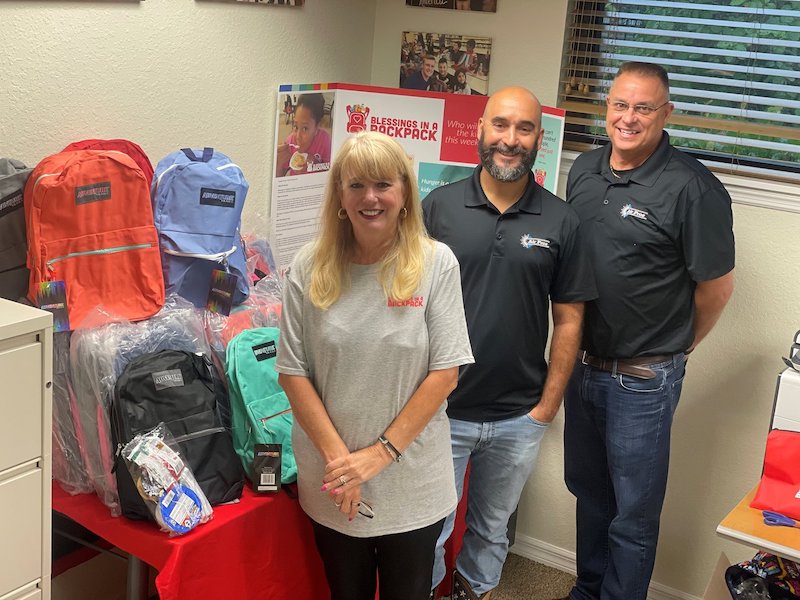 Three adults stand beside a table filled with colorful backpacks and supplies, supporting food insecure students in front of a "Blessings in a Backpack" display at the Air Pros USA office.