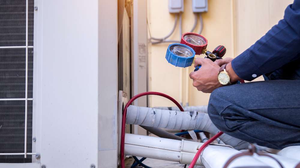A person checks a central air conditioning unit for issues using pressure gauges, with wires and pipes visible around the equipment.