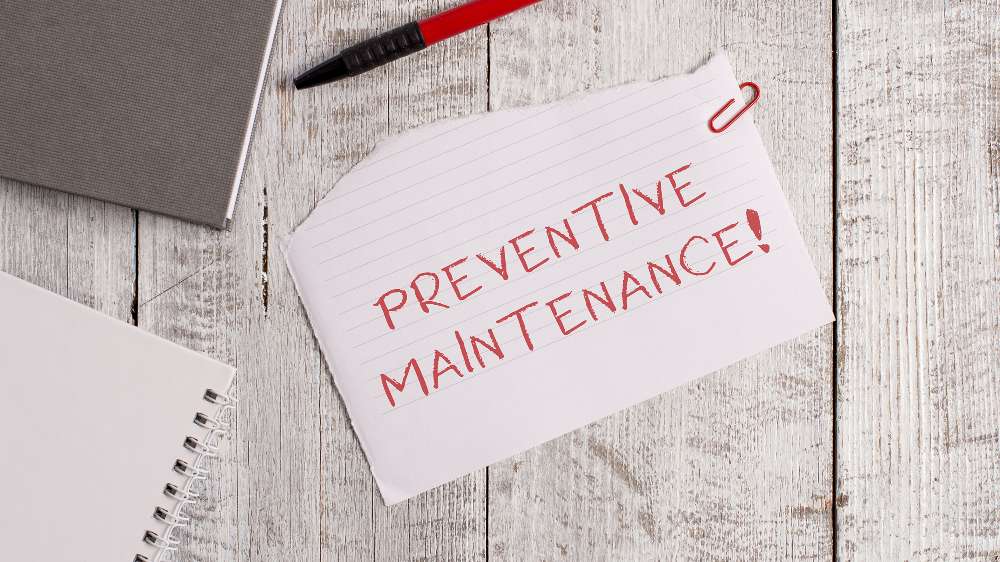 A piece of lined paper with "PREVENTIVE MAINTENANCE!" written in red marker, clipped with a red paperclip, placed on a wooden surface next to a pen and notebooks—reminding you to extend your central AC lifespan.
