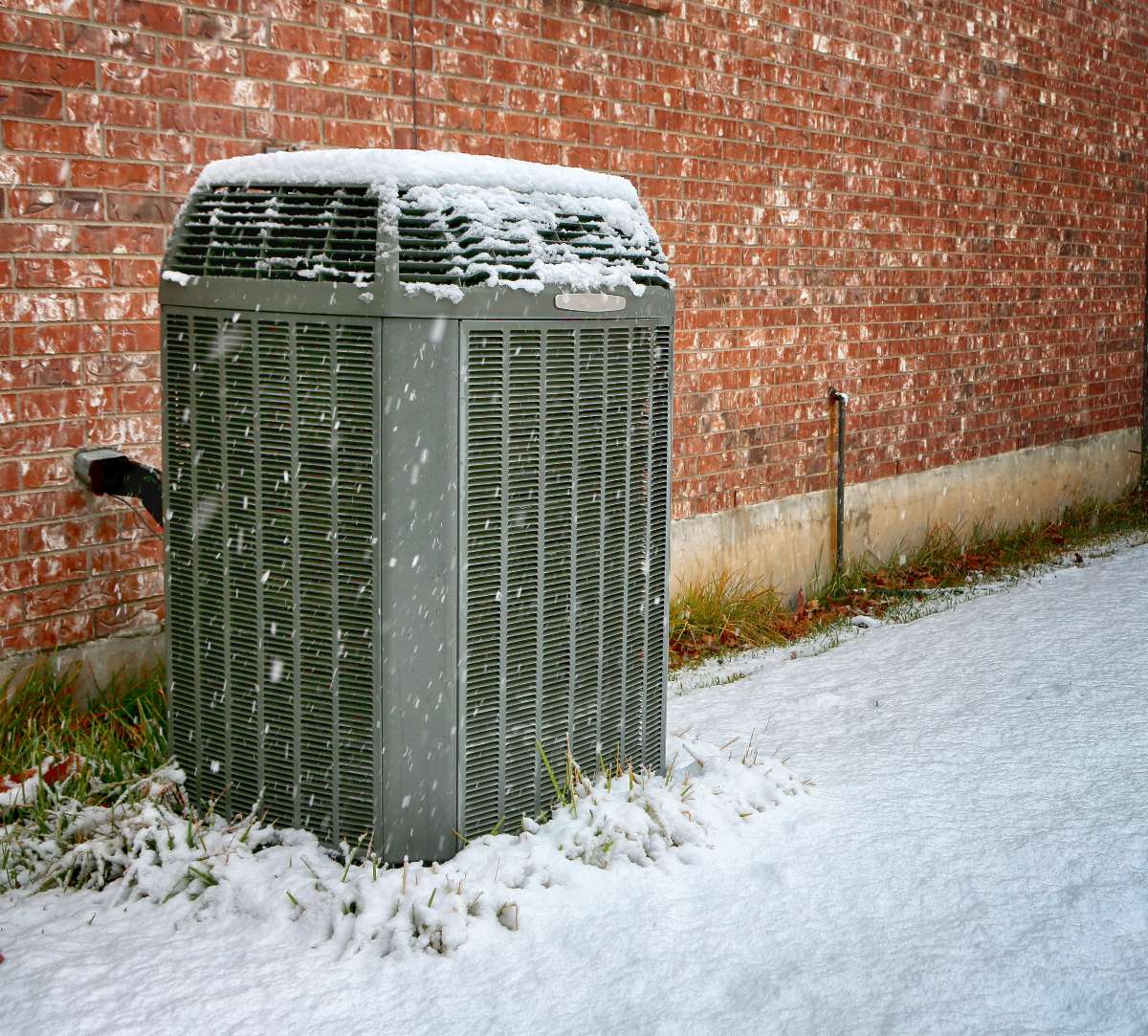 A central air conditioning unit outdoors, partially covered with snow, sits next to a red brick building with a snow-covered ground—reminding you that winter is the best time to schedule AC services for the upcoming warmer months.