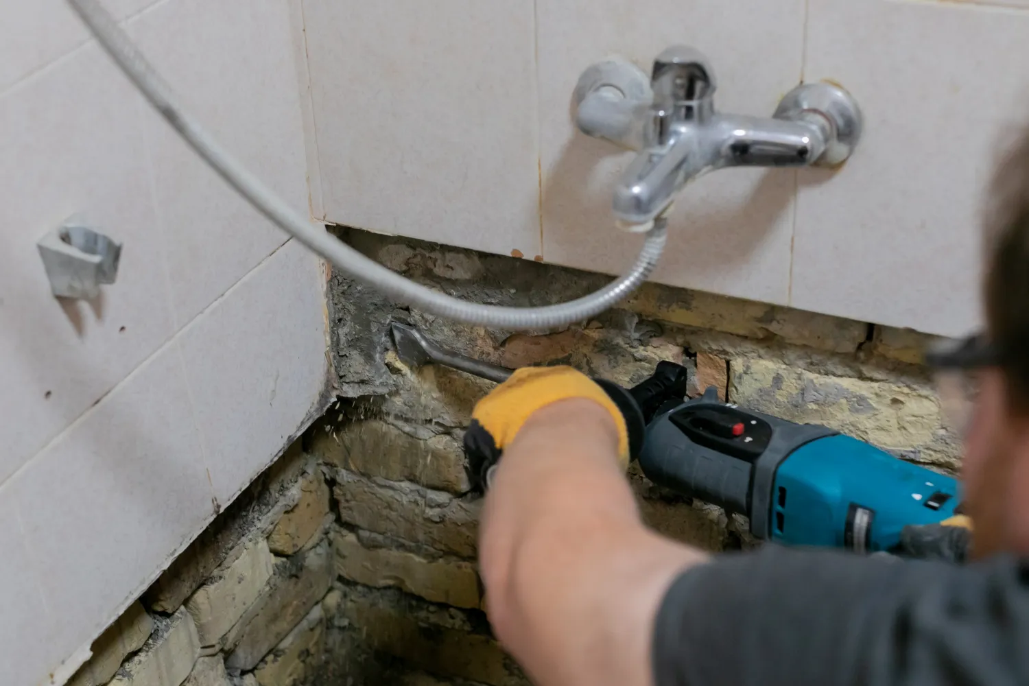 A person wearing yellow gloves uses an electric drill to remove tiles and plaster from a wall under a bathroom faucet, revealing brickwork underneath.