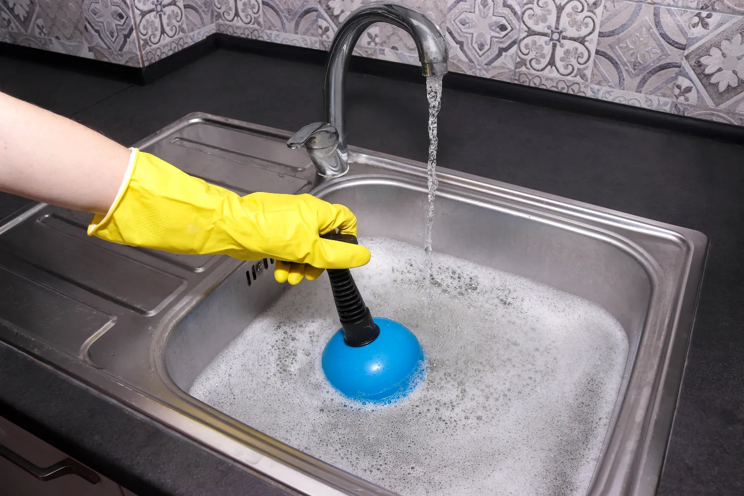 A person wearing a yellow glove uses a blue plunger in a kitchen sink filled with soapy water under a running faucet, possibly performing sink repair.