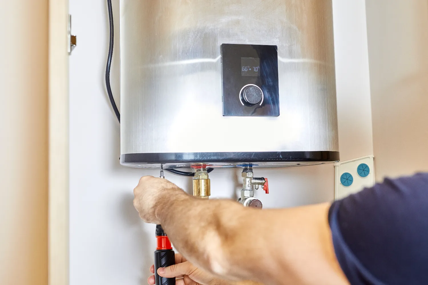 Person using a screwdriver to tighten a screw at the bottom of a wall-mounted stainless steel water heater during water heater repair.