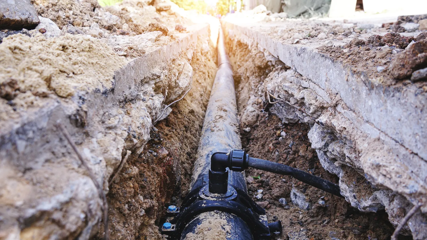 A large black pipe for water line installation is set in a trench dug through soil and concrete, with sunlight visible at the end of the trench.
