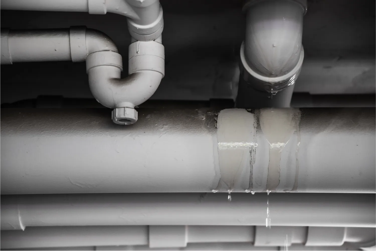 A close-up view of gray pipes on a ceiling, with water visibly leaking and dripping from a joint—an urgent sign that water line repair is needed.
