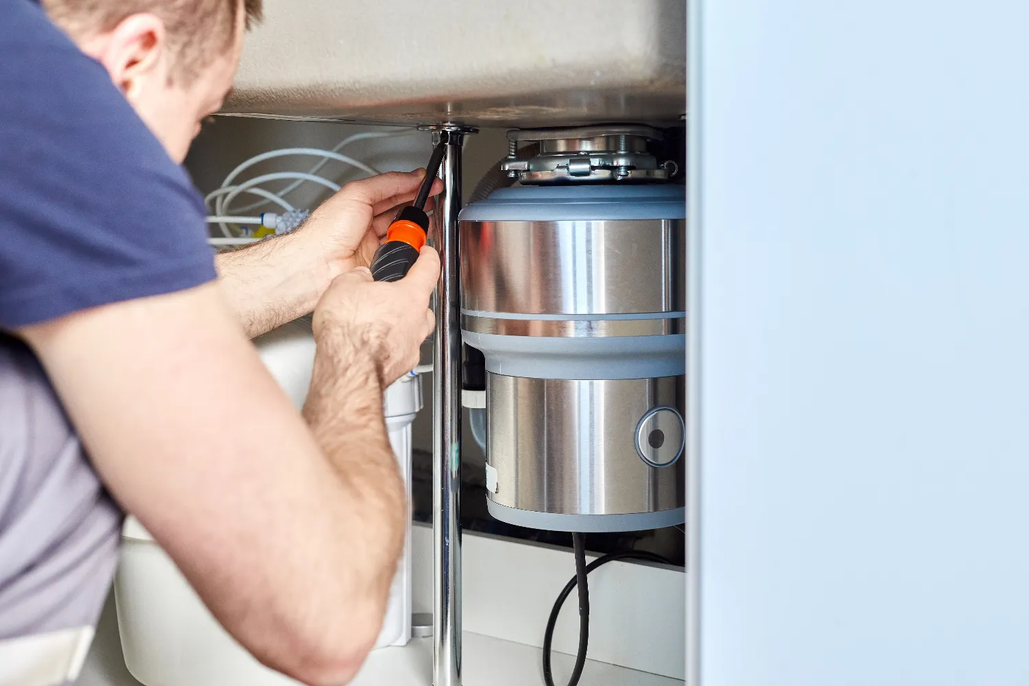 A person uses a screwdriver to install or repair a garbage disposal beneath the kitchen sink.
