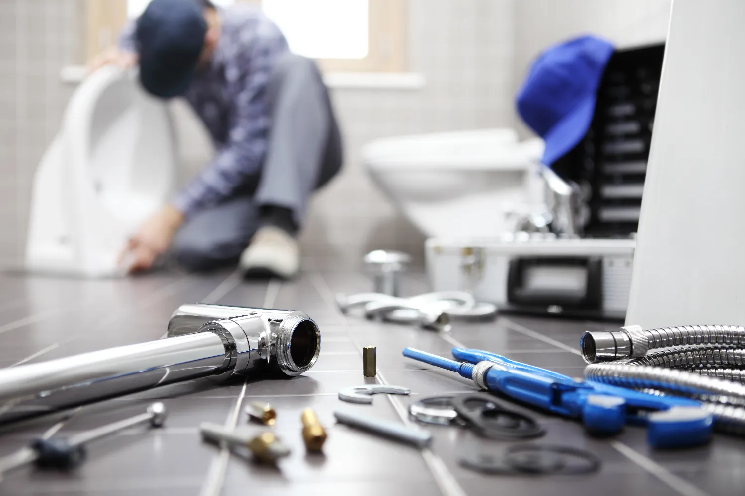 A plumber performs a toilet repair in a bathroom, with plumbing tools and parts for toilet installation spread out on the tiled floor in the foreground.