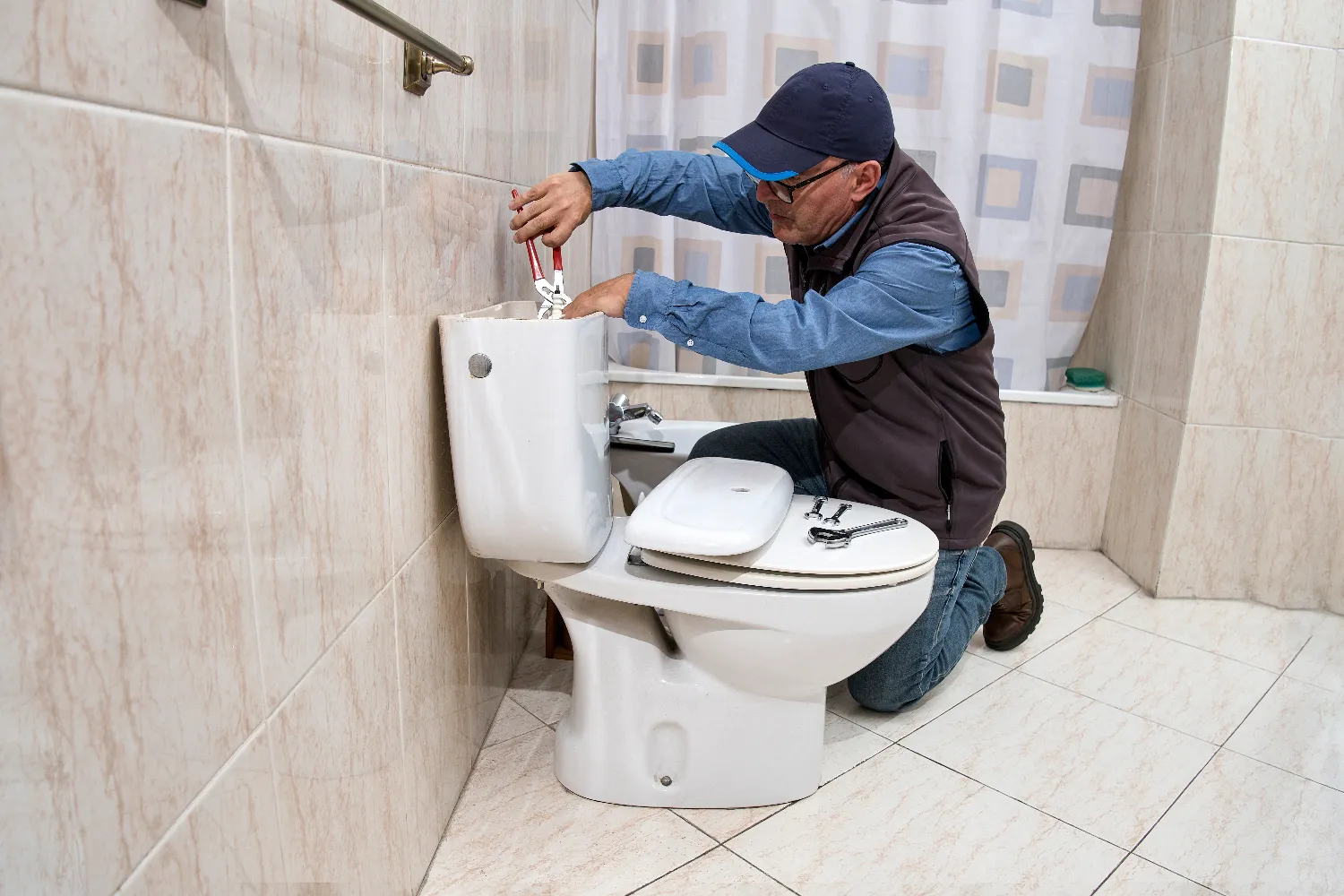 A plumber wearing a cap and glasses uses a wrench for toilet repair, fixing the tank in a tiled bathroom.