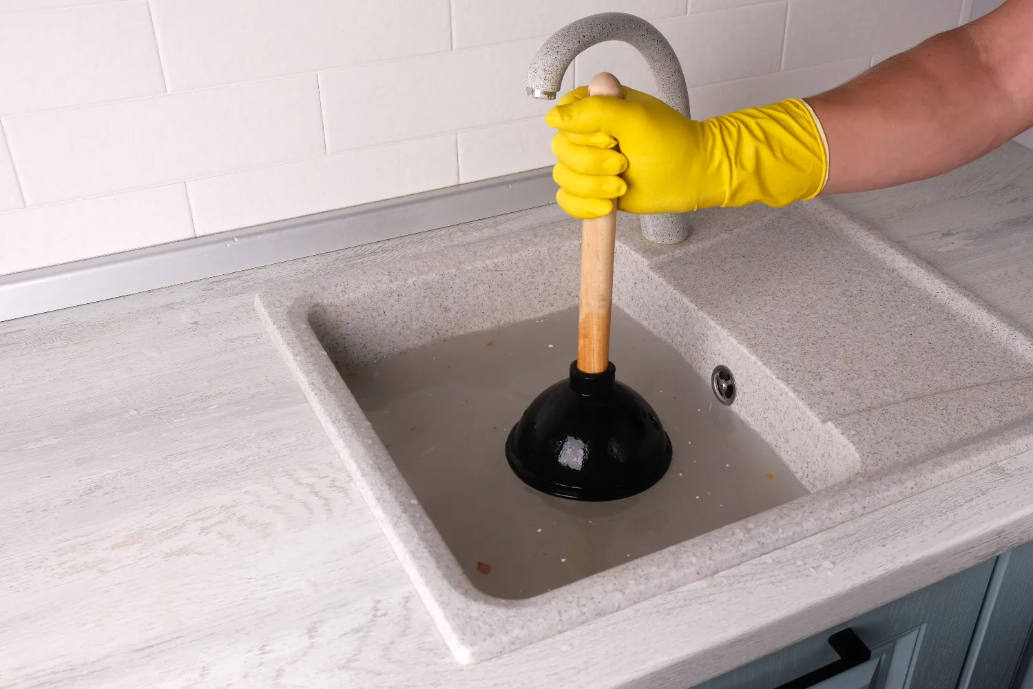 A person wearing a yellow glove uses a plunger for drain cleaning to unclog a kitchen sink filled with water.